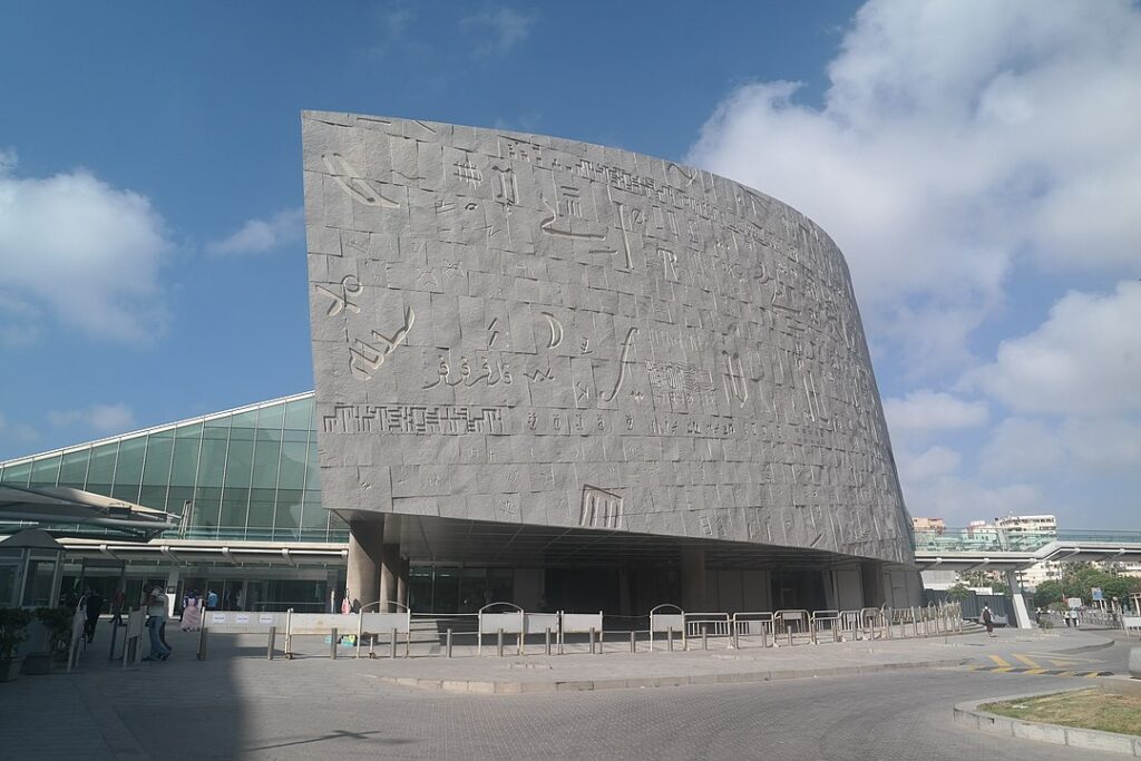 新亚历山大图书馆（Bibliotheca Alexandrina）的外观，于2002年开放。其创新的圆盘形设计唤起了古埃及太阳神的形象，象征着古代智慧与现代技术的融合。这座宏伟的海滨建筑作为现代知识殿堂，承载了古代图书馆的遗产，吸引了全球的关注。